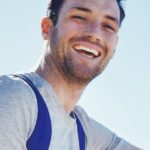 Ein junger Mann mit grauem Shirt und blauem Rucksack, der entspannt lacht