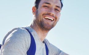 Ein junger Mann mit grauem Shirt und blauem Rucksack, der entspannt lacht