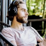 Junger Mann sitzt auf der Veranda einer Waldhütte. Er hat Kopfhörer auf und blickt in die Ferne.