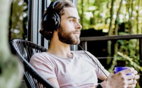 Junger Mann sitzt auf der Veranda einer Waldhütte. Er hat Kopfhörer auf und blickt in die Ferne.