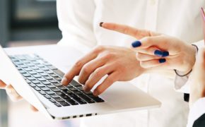 Zwei Frauen im Businessdress in Nahaufnahme. Man sieht nur die Hände. Die eine hält einen Laptop und schreibt auf, was ihr die andere diktiert. Die Zweite hat blau lackierte Fingernägel und den den Zeigefinger in Richtung des Laptop Displays ausgestreckt.