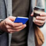 Mann mit Jacket, einem Handy in der einen und einem Kaffeebecher in der anderen Hand.