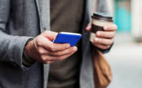 Mann mit Jacket, einem Handy in der einen und einem Kaffeebecher in der anderen Hand.
