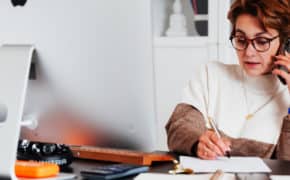Frau sitzt am Schreibtisch, telefoniert und schreibt etwas auf ein Blatt Papier.