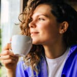 Junge Frau sitzt im Café mit Tasse in der Hand und schaut verträumt aus dem Fenster.