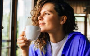 Junge Frau sitzt im Café mit Tasse in der Hand und schaut verträumt aus dem Fenster.