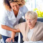 Eine Pflegerin hilft einer älteren Frau aus einem Sessel.
