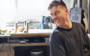 Lachender blonder junger Mann sitz in einer Werkstadt und schaut nach hinten.