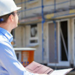 Ein Mann steht auf einen Bauplan schauend auf ein Baustelle von einem Haus.