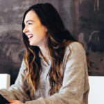 Eine junge Frau mit einem Tablet in der Hand sitzt an einem Konferenztisch in einem Konferenzraum und lacht.
