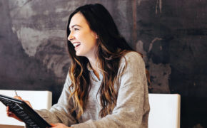 Eine junge Frau mit einem Tablet in der Hand sitzt an einem Konferenztisch in einem Konferenzraum und lacht.
