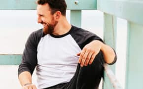 Lachender Mann sitzt auf einem Balkon und schaut nach rechts.