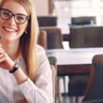 Frau mit Brille und Stift sitzt am Tisch und lacht