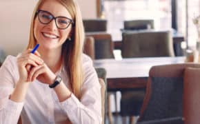 Frau mit Brille und Stift sitzt am Tisch und lacht