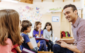 Ein Lehrer sitzt vor einer Gruppe an Kindern.