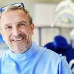 Ein Zahnarzt schaut lächelnd in die Kamera, eine Zahnarzthelferin bereitet die Geräte im Hintergrund vor.