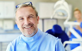 Ein Zahnarzt schaut lächelnd in die Kamera, eine Zahnarzthelferin bereitet die Geräte im Hintergrund vor.
