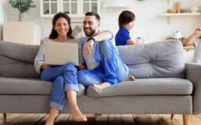 Ein Paar sitz auf einem Sofa und bespricht etwas an ihrem Laptop während ihre Kinder spielen.
