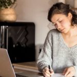 Private Krankenversicherung für Selbstständige und Freiberufler. Eine Frau sitzt zu Hause am Schreibstisch vor ihrem Laptop und schreibt etwas mit der Hand auf ein Blatt Papier.