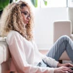 Eine Frau sitzt vor ihrer Couch und tippt auf ihrem Laptop.