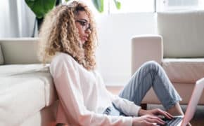 Eine Frau sitzt vor ihrer Couch und tippt auf ihrem Laptop.