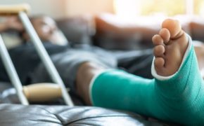 Ein Mann liegt auf dem Sofa, eine Krücke neben sich. Bein und Fuß sind eingegipst und mit einem grünen Verband umwickelt.