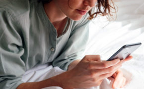 Eine brünette Frau liegt auf dem Bett und schaut auf ihr Smartphone.