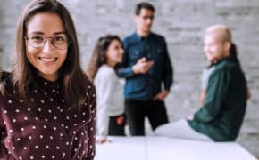 Frau im Meetingraum vor einem Tisch. Hinten unterhalten sich drei junge Angestellte. Frau guckt in die Kamera