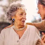 Eine alte Frau im Großmutteralter, die von einer jüngeren Frau, wahrscheinlich die Tochter, in den Arm genommen wird. Mit ihrer linken Hand drückt sie die Hand der alten Frau.