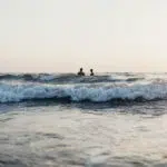 zwei Badende im Meer, fotografiert vom Strand vor anrauschenden Brandung und bei Sonnenuntergang. Wie man sich diesen Traum mit einer Reiserücktrittversicherung auch gegen Corona bewahren kann