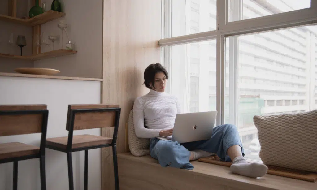 Eine junge Frau sitzt in einer Fensternische auf einer Fensterbank. Durch das Fenster erkennt man im Hintergrund ein Hochhaus. Die Frau ist an die Wand gelehnt und tippt in ihr MacBook auf ihrem Schoß 