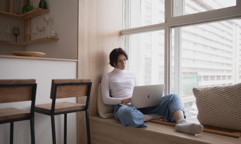 Eine junge Frau sitzt in einer Fensternische auf einer Fensterbank. Durch das Fenster erkennt man im Hintergrund ein Hochhaus. Die Frau ist an die Wand gelehnt und tippt in ihr MacBook auf ihrem Schoß